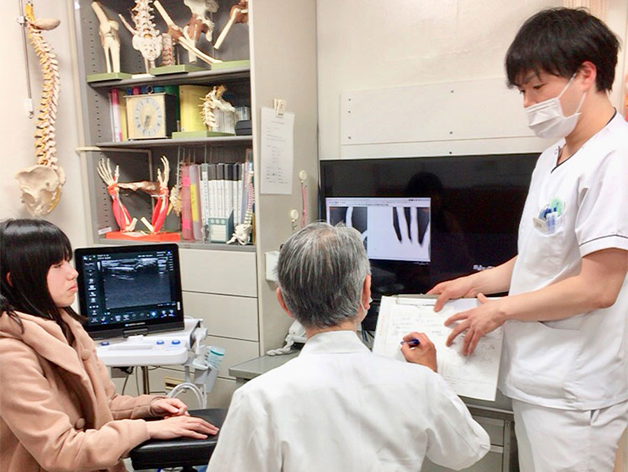 整形外科の診察室で、超音波画像やX線画像を前に医師と打ち合わせる院長