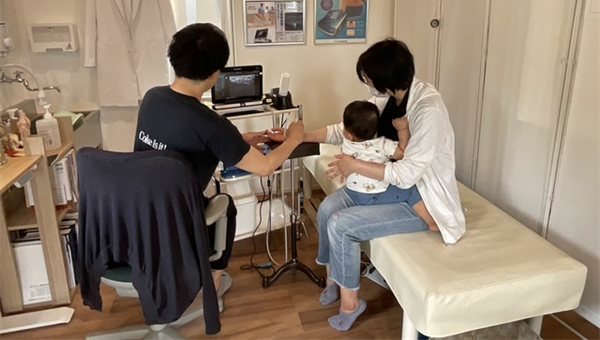 施術室で、母親に抱かれた乳児の腕にエコーを当て、画面で状態を確かめる院長