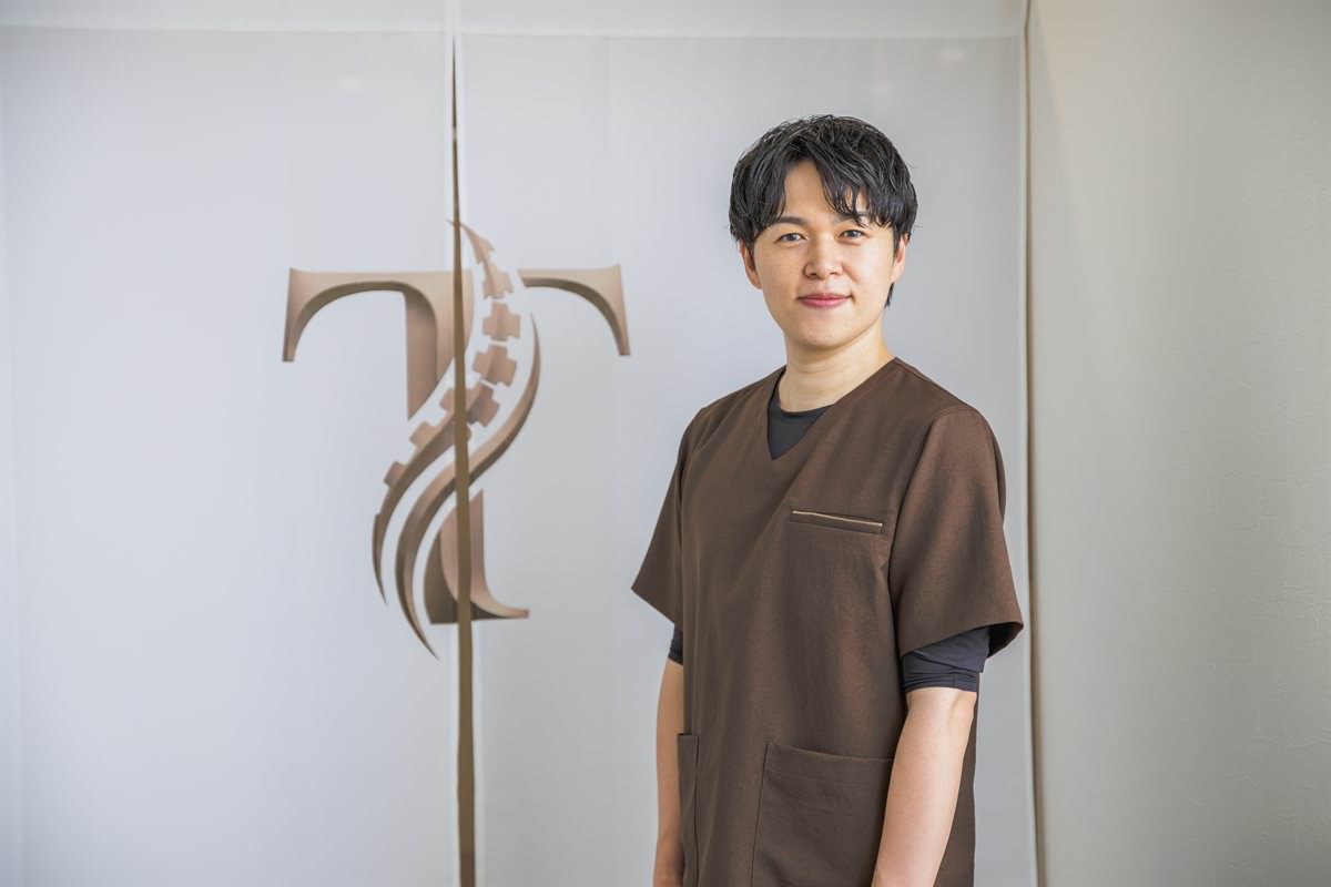 院のロゴが入ったのれんの前に立つ院長・髙野正和
