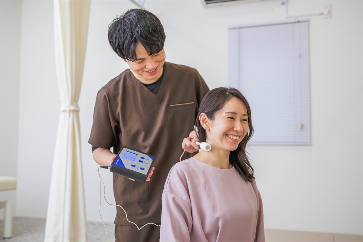 微弱電流の機器NEUBOXを手に、笑顔の患者さまの首まわりに電極を当てる院長