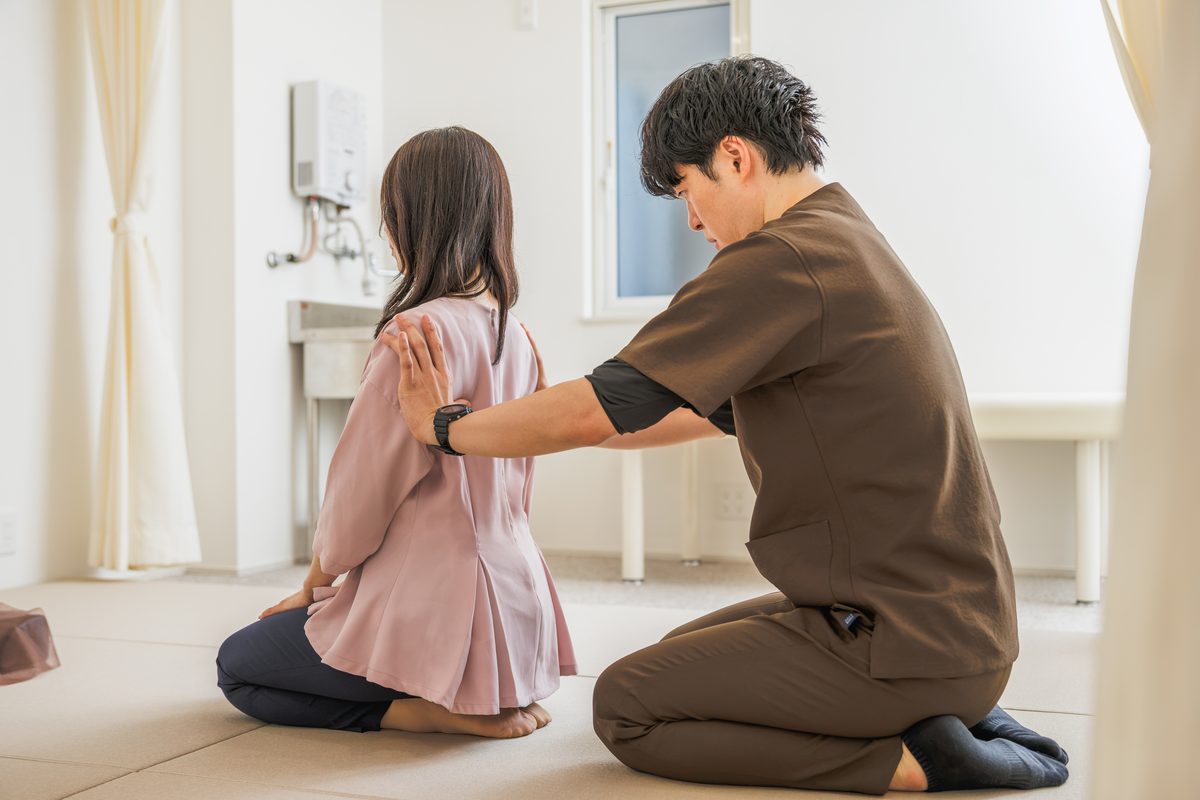 畳に正座した患者さまの後ろにひざをつき、背中と腕に手を当てて体を整える院長