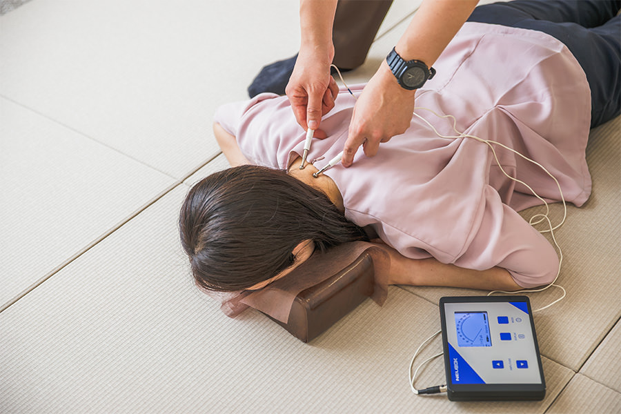 畳の上でうつ伏せになった患者さまの肩の一点に、細い電極を当ててトリガーポイント療法をおこなう院長