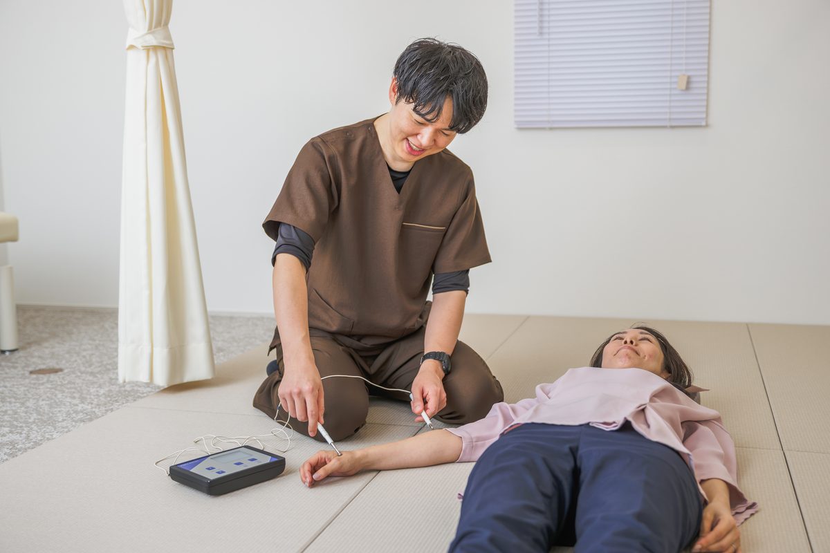 畳の上で仰向けになった患者さまの手首に、ペン型の電極を当てる院長